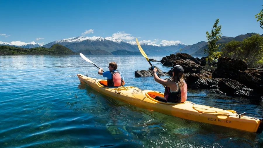 Two people paddling a double kayak near the shore of Lake Wanaka with mountain views.