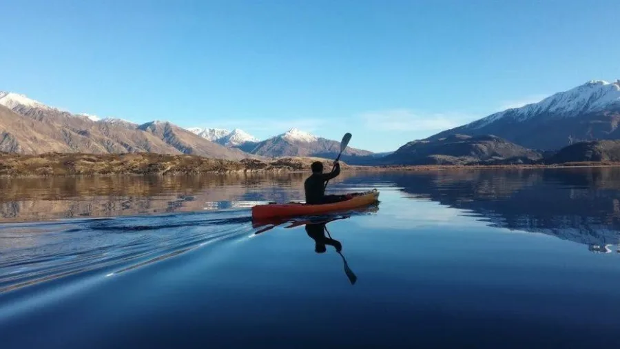 Person kayaking solo on a calm Lake Wanaka with snow-capped mountains in the background.