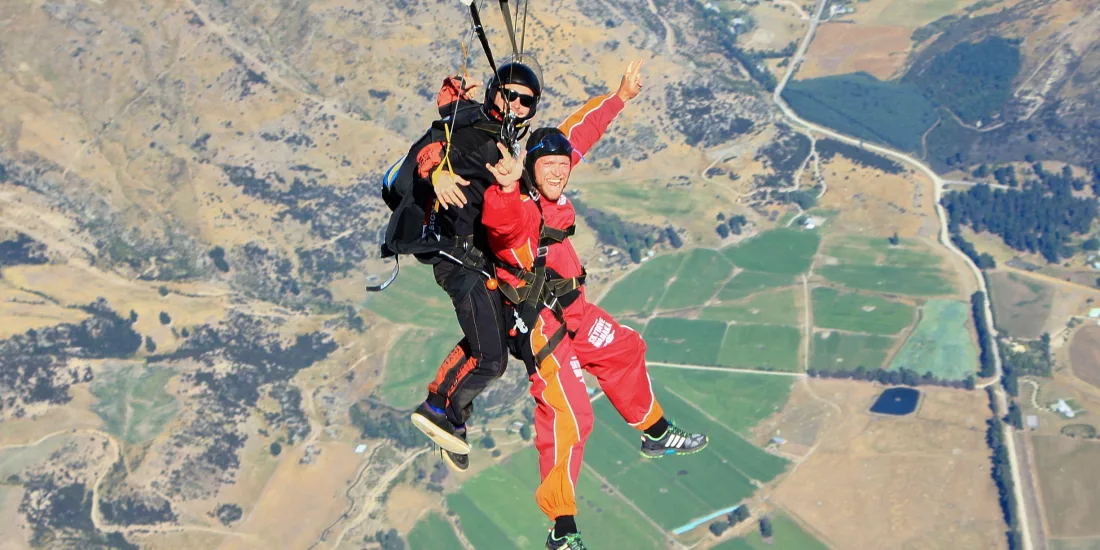 Tandem skydivers gliding over farmland near Wanaka under a green parachute