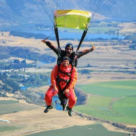 Two tandem skydivers mid-air above Wanaka farmland smiling at the camera