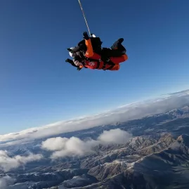 Two tandem skydivers freefalling above the Southern Alps with mountain ranges and clouds beneath