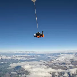 Tandem skydiver falling with parachute line extended high above Otago and scattered cloud