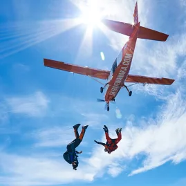 Two pairs of skydivers falling under bright sun beside Skydive Wanaka aircraft