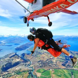 Skydivers leaping from plane with aerial view of farmland and Lake Wanaka in distance