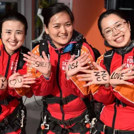 Three smiling skydivers in jumpsuits showing hand-written messages on their palms