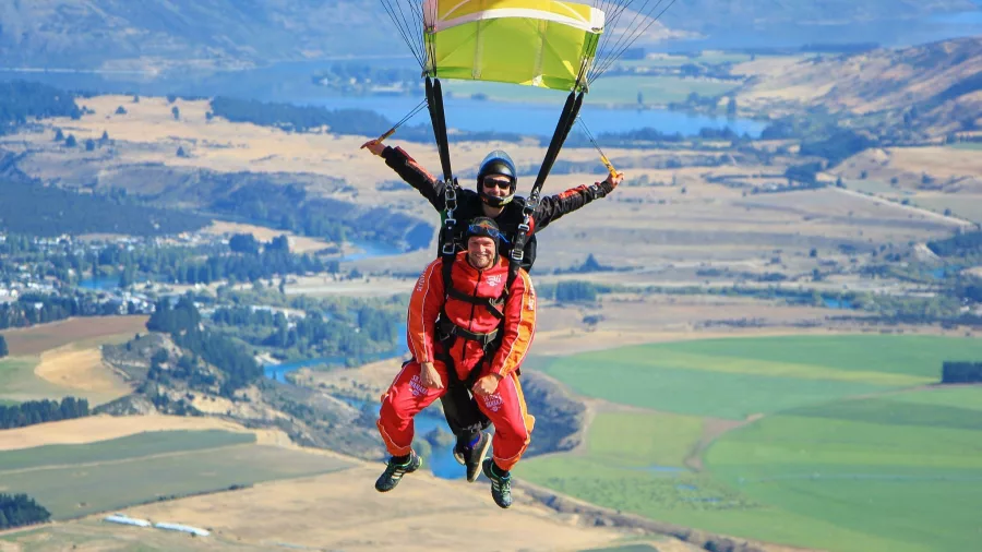 Two tandem skydivers mid-air above Wanaka farmland smiling at the camera