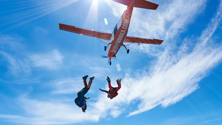 Two pairs of skydivers falling under bright sun beside Skydive Wanaka aircraft