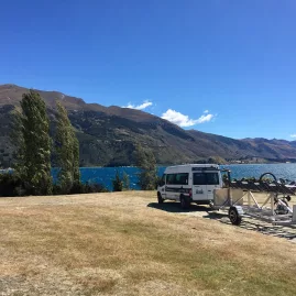 Bike trailer and support van parked near Lake Hawea with lake and mountains in background