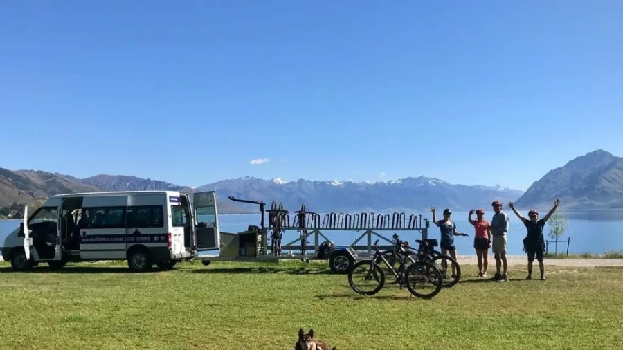 Cyclists waving beside shuttle van and bikes at Lake Hawea with a dog relaxing in foreground