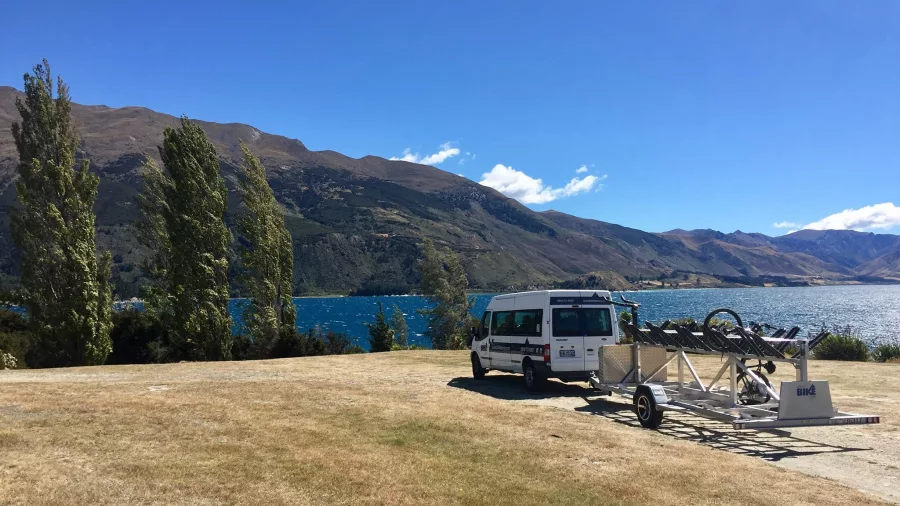 Bike trailer and support van parked near Lake Hawea with lake and mountains in background