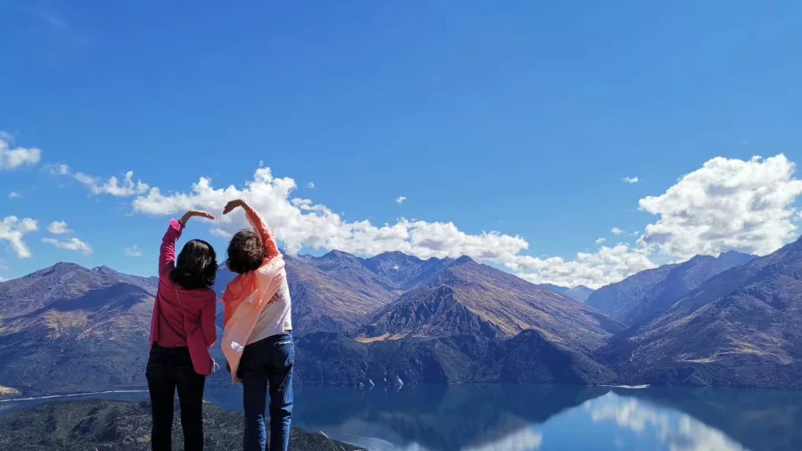 Two people making heart shapes with their arms overlooking Lake Wanaka and the mountains.