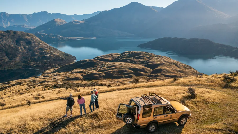 Group standing near a 4WD vehicle overlooking Lake Wanaka and alpine terrain.