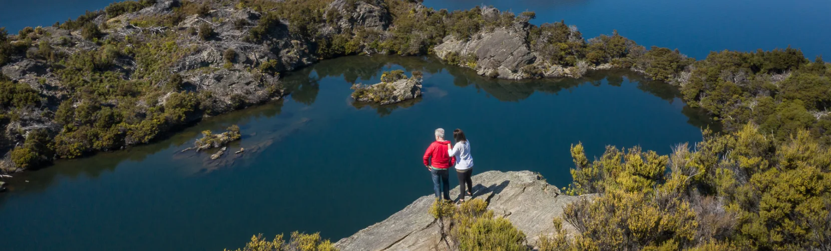 A couple stands at the top of Mou Waho Island, looking out over Arethusa Pool and Lake Wanaka.