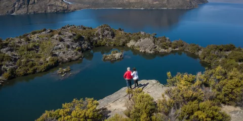 A couple stands at the top of Mou Waho Island, looking out over Arethusa Pool and Lake Wanaka.