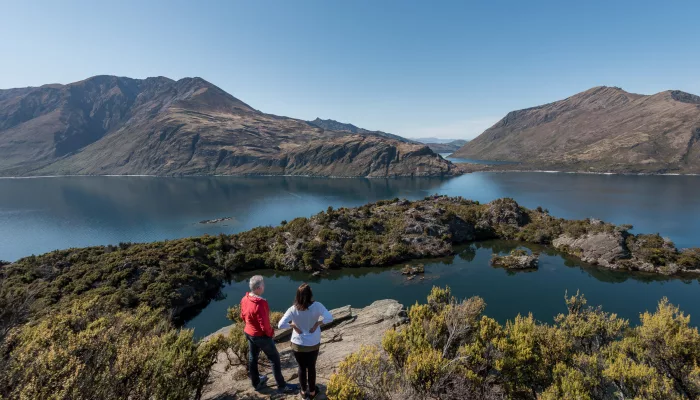 A couple stands at a lookout above Arethusa Pool on Mou Waho Island, taking in the expansive lake views.