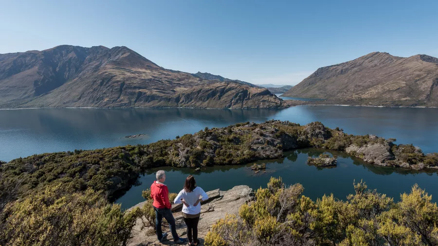 A couple stands at a lookout above Arethusa Pool on Mou Waho Island, taking in the expansive lake views.