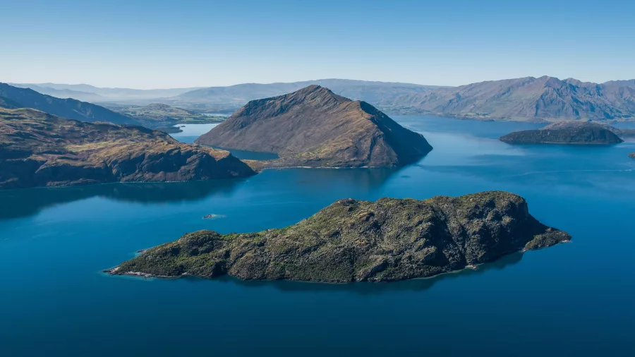 An aerial panorama of Mou Waho Island surrounded by Lake Wanaka, with rugged mountains and valleys beyond.