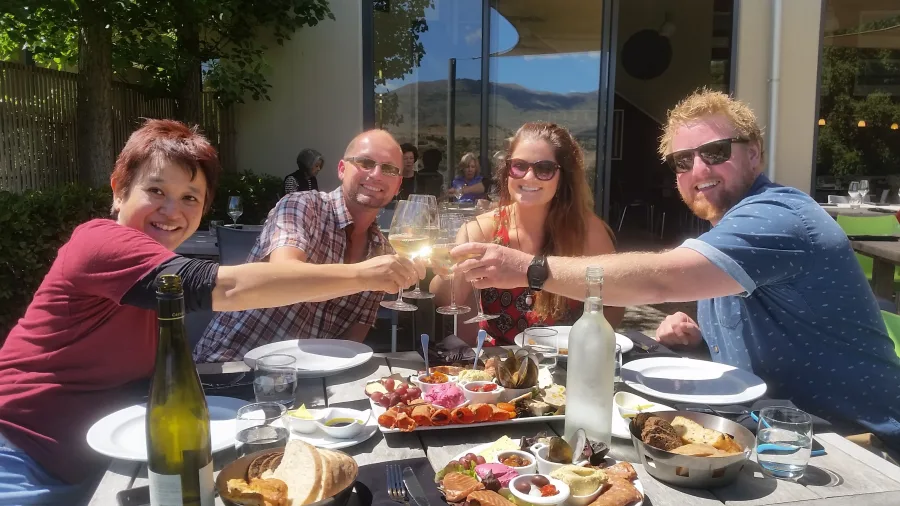 A cheerful group toasts glasses of white wine over a vineyard lunch spread on a sunny afternoon in Wanaka.