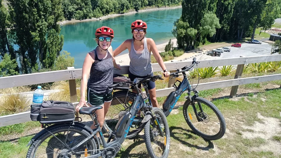 Two smiling cyclists at the Clyde end of the Lake Dunstan Trail with river views.