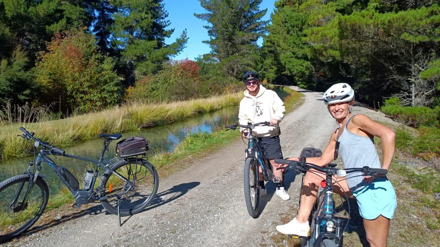 Two cyclists taking a break beside a calm section of the Hawea River Trail near Wanaka.
