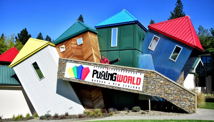 Exterior of Puzzling World in Wānaka featuring colourful tilted buildings and a visitor posing on the ramp