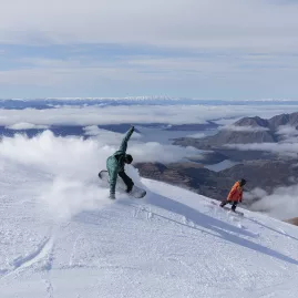 Snowboarders carving down the slopes of Treble Cone with panoramic views over Lake Wānaka in New Zealand.