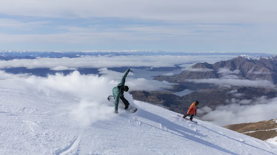 Snowboarders carving down the slopes of Treble Cone with panoramic views over Lake Wānaka in New Zealand.