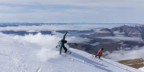 Snowboarders carving down the slopes of Treble Cone with panoramic views over Lake Wānaka in New Zealand.