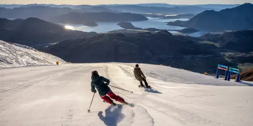 Two skiers carving down a snow-covered slope at Treble Cone Ski Area with panoramic views of Lake Wānaka and the Southern Alps