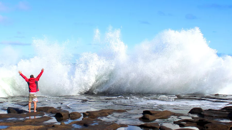 Person in red jacket standing on rocky shore as powerful wave crashes at Curio Bay on the Southland coast in the Catlins, New Zealand