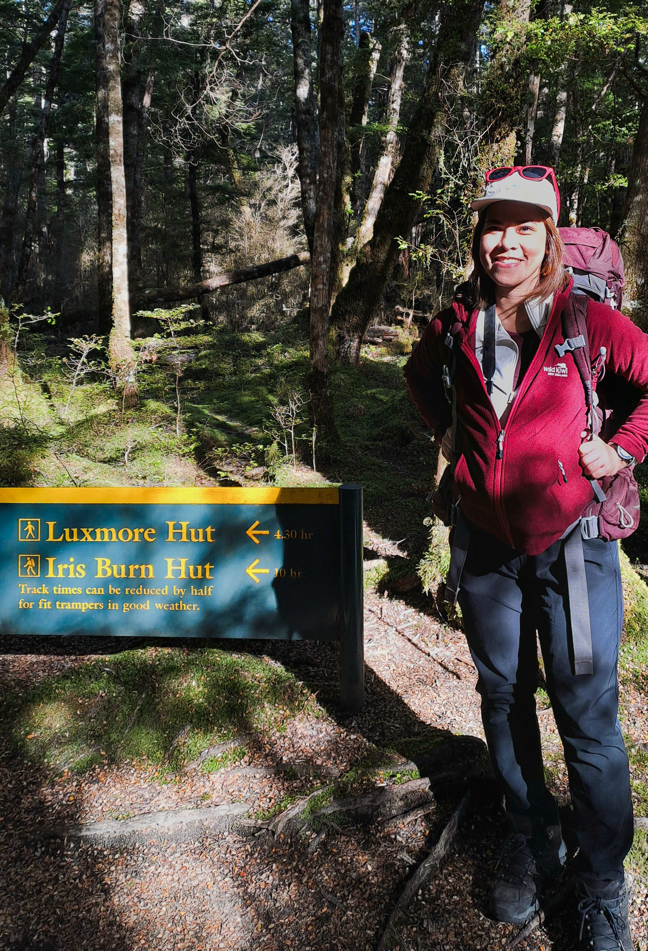 Jen stands beside a green and yellow DOC sign indicating the track times for Luxmore Hut and Iris Burn Hut on the Kepler Track.