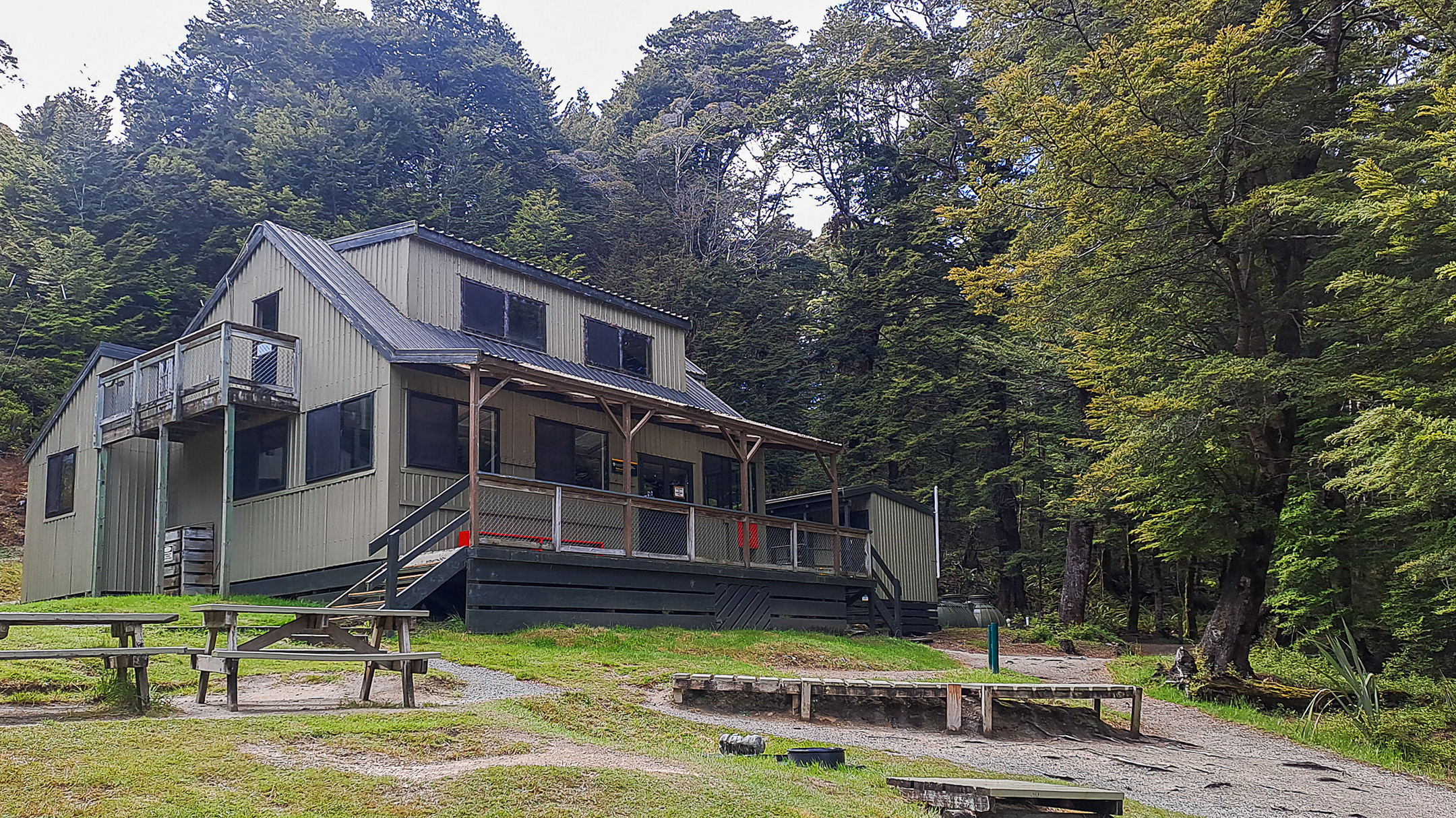 A two-storey wooden building with a balcony and picnic tables stands in a forest clearing on the Kepler Track.