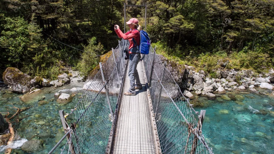 Hiker photographing from swing bridge over turquoise river on Lake Marian Track, Southland New Zealand