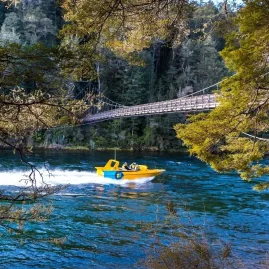 Jet boat cruising under a suspension bridge surrounded by native forest on the Waiau River.