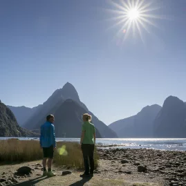 Two people admire Mitre Peak from the Milford Sound foreshore under a clear blue sky.