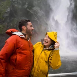 Couple laughing in colourful rain jackets while getting sprayed near Stirling Falls on a Milford Sound nature cruise.