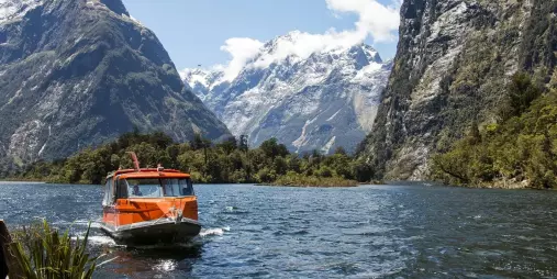 Water taxi arrives at Sandfly Point with snow-capped peaks rising behind in Milford Sound.