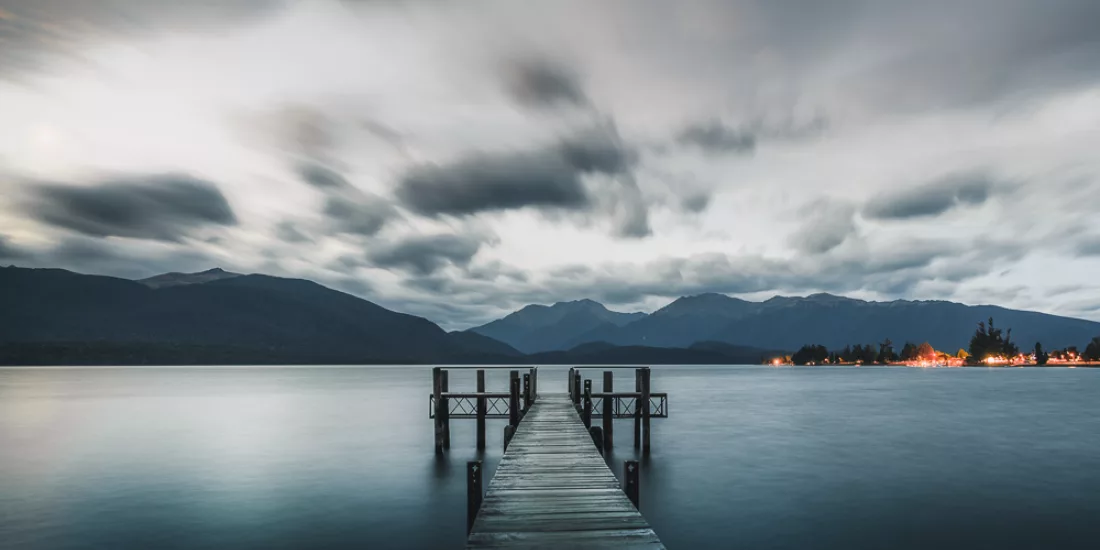 Wooden wharf stretching into the still waters of Lake Te Anau at sunset with dramatic clouds and Fiordland mountains in the background