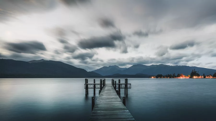 Wooden wharf stretching into the still waters of Lake Te Anau at sunset with dramatic clouds and Fiordland mountains in the background