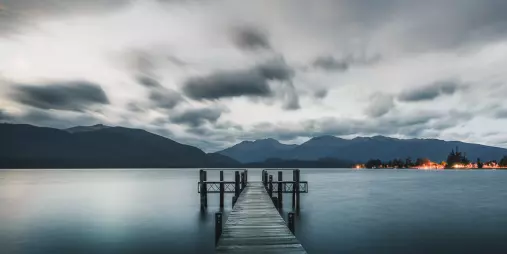 Wooden wharf stretching into the still waters of Lake Te Anau at sunset with dramatic clouds and Fiordland mountains in the background
