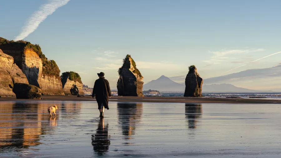 Woman walking her dog on Tongaporutu Beach beside the Three Sisters sea stacks with Mount Taranaki in the distance