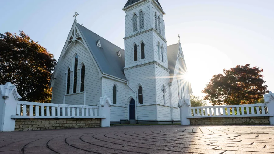 St. Andrews Anglican Church in Cambridge, Waikato, New Zealand, with its white wooden exterior, tall steeple, and sunrise light shining through the trees.