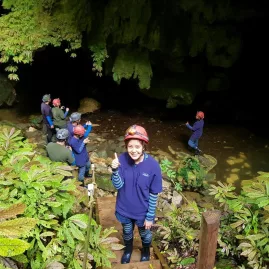 Visitors wearing helmets and outdoor gear gather at the forested entrance to a cave on a guided Waitomo tour.