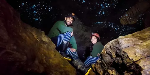 Smiling couple in caving gear sitting beneath a glow worm-lit ceiling inside a Waitomo cave.