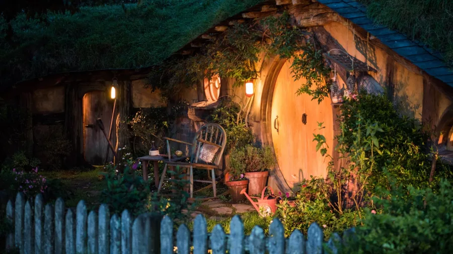 Cosy hobbit hole at Hobbiton Movie Set lit by lanterns at dusk with garden and picket fence