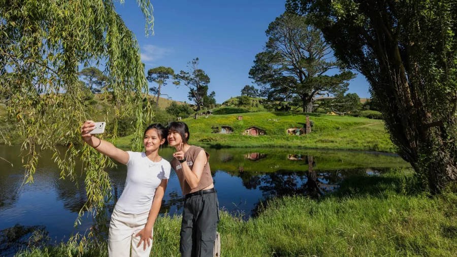 Two visitors taking a selfie near the Hobbiton lake with Hobbit Holes in the background
