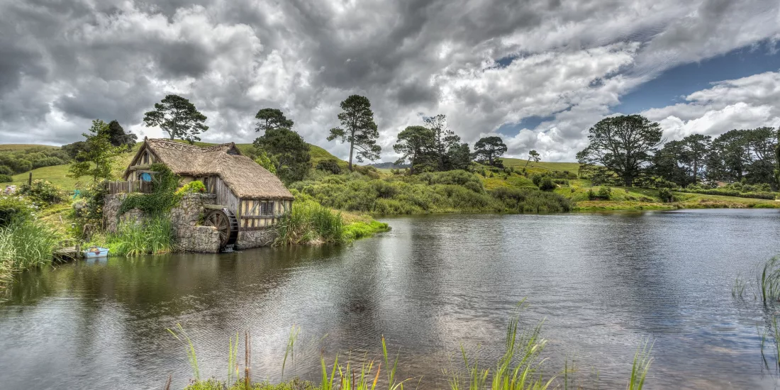 Thatched mill building beside the lake at Hobbiton Movie Set, with trees reflected in the still water