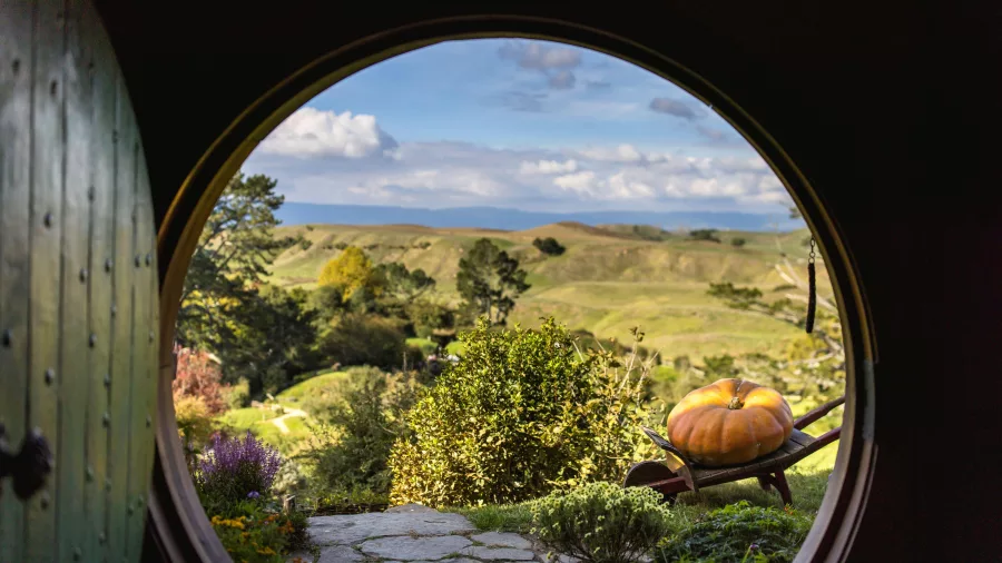View through round hobbit door at Hobbiton Movie Set with wheelbarrow and pumpkin overlooking Matamata hills