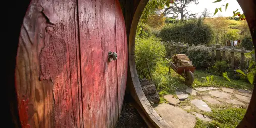 View from inside a hobbit hole at Hobbiton Movie Set looking out through a red round door onto a sunny garden in Matamata