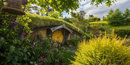 Sunlight peeking through trees onto a yellow-door Hobbit hole surrounded by a lush garden at Hobbiton Movie Set